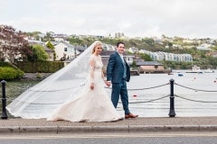 Bride and groom walking on harbour edge with veil trailing behind
