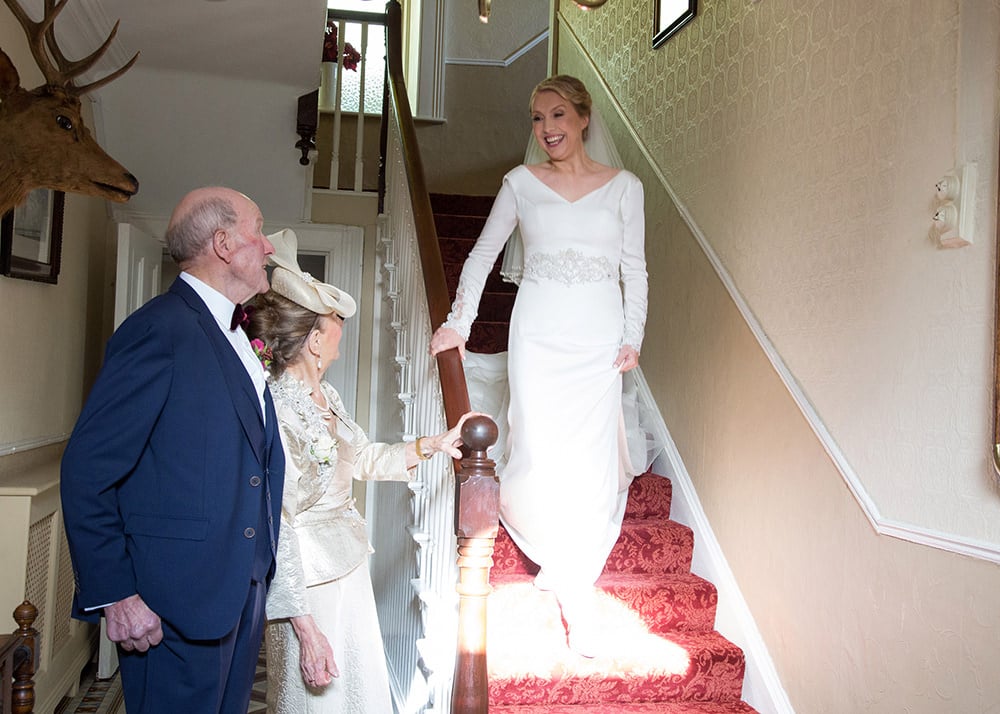 Bride descends the stairs while her parents wait at the bottom to greet her
