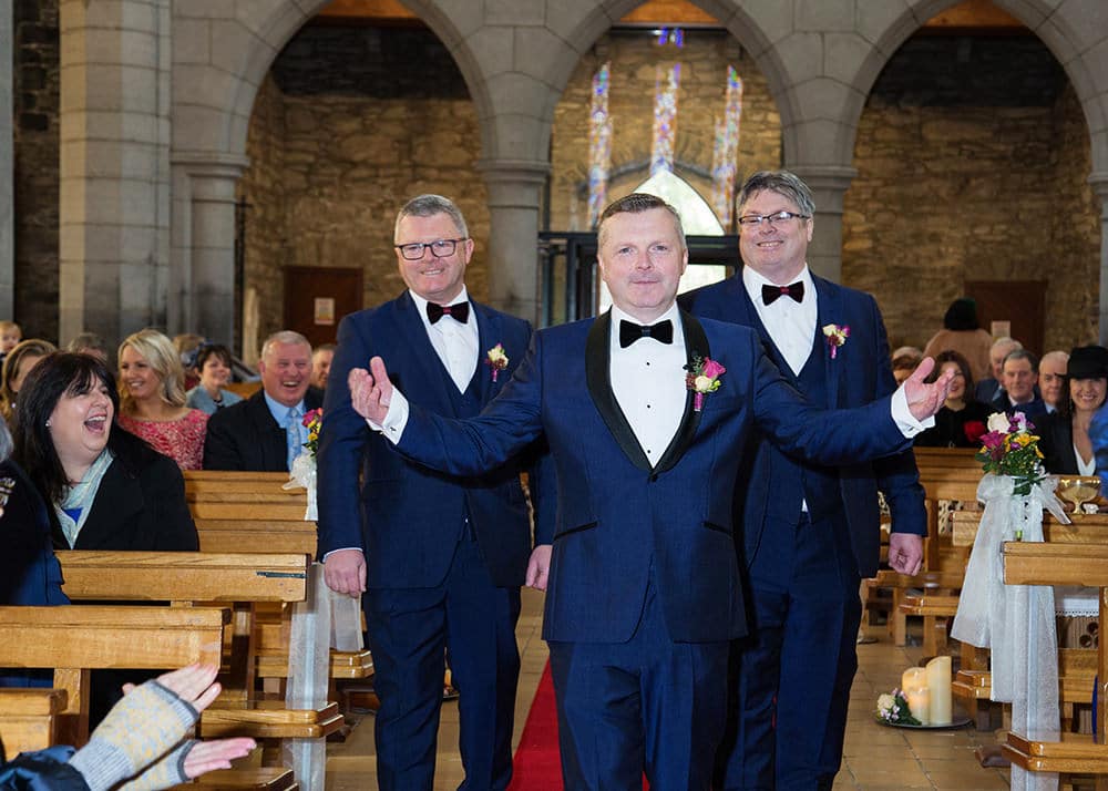 Groom and groomsmen smile from the aisle of the church while guests look on
